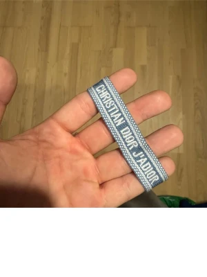 Blått Christian Dior J'Adior armband - Snyggt blått och vitt vävt armband från Christian Dior med texten 'J'Adior'. Armbandet har tofsar i ändarna och är gjort i textilmaterial. Perfekt accessoar för att lyfta din outfit med en exklusiv touch.