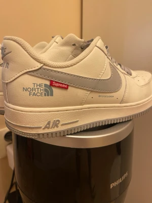 Vita sneakers Air x Supreme x TNF - Unika vita sneakers med grå swoosh från Nike Air, dekorerade med The North Face och Supreme-logga på sidan. Skon har lågt skaft och är tillverkad i slätt läder med klassisk Air-sula. Perfekt för dig som gillar streetwear och samarbeten.