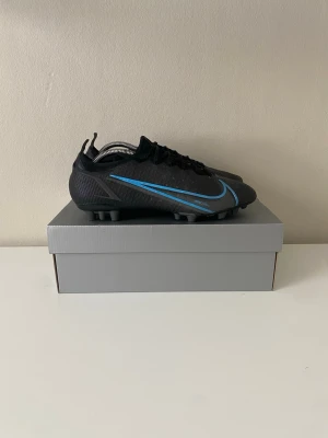 Nike Mercurial svart/blå fotbollsskor Elite  - Säljer ett par Nike Mercurial fotbollsskor i svart med blå detaljer och klassisk Nike-swoosh på sidan. Skorna har snörning, låg profil och dobbar för bra grepp på konstgräs men fungerar även på gräs.  Säljer för att dem är för små.