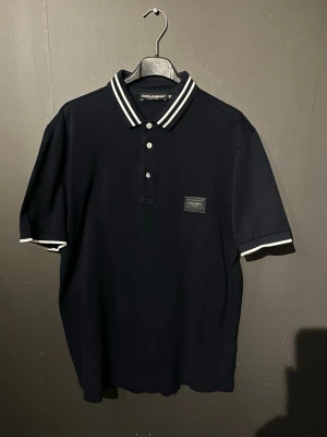 Svart piké från Dolce & Gabbana - Klassisk svart pikétröja från Dolce & Gabbana med vitrandiga detaljer på krage och ärmslut. Tröjan har korta ärmar, knappar framtill och en diskret logopatch på bröstet. Perfekt för en clean och stilren look. (Intressekoll) dma vid intresse!