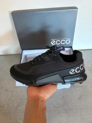 Svarta sneakers från ECCO - Snygga svarta sneakers från ECCO med mesh- och syntetmaterial, coola reflexdetaljer på snörena och stor logga på sidan. Skorna har en chunky sula och modern design som passar perfekt till streetwear. Låg modell med bekväm passform.