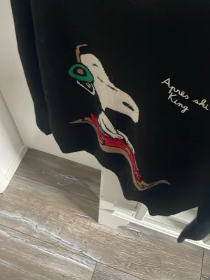 Svart stickad tröja MC2 St. Barth -    Knappt använd Cool svart stickad tröja från MC2 St. Barth Après Ski Collection med en stor Snoopy-figur på framsidan och texten 'Après ski king'. Tröjan har färgglada detaljer i rött, grönt och vitt. Perfekt för dig som gillar statement-plagg och snygga prints.