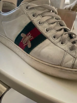 Gucci vita sneakers med bi och grönt skinn - Säljer ett par vita Gucci sneakers med klassiska gröna och röda ränder på sidan samt en broderad guldfärgad bi. Baksidan har en detalj i grönt ormskinnsmönster. Skorna har lågt skaft och är i läder med textilinslag.