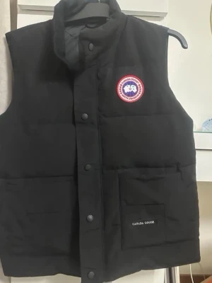 Svart dunväst från Canada Goose - Snygg svart dunväst från Canada Goose med hög krage och tryckknappar framtill. Västen har en tydlig logotyp på bröstet och praktiska fickor med lock. Tillverkad i slitstarkt material och fylld med dun för extra värme. Perfekt för kalla dagar.