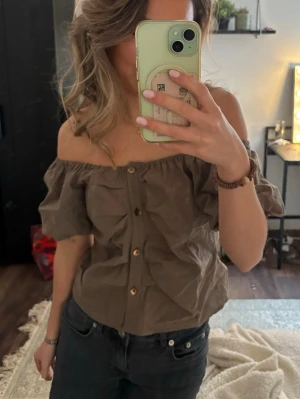 Brun offshoulder topp med knappar - Snygg brun offshoulder topp med korta puffärmar och knappar framtill. Toppen har en avslappnad passform och är perfekt för dig som vill ha en trendig och luftig look. Tillverkad i bomull för en skön känsla.