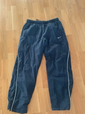Marinblå träningsbyxor från Nike - Säljer ett par marinblå träningsbyxor från Nike med vit logga på vänster ben och vita detaljer längs sidorna. Byxorna har resår i midjan och är tillverkade i ett lätt, sportigt material som passar perfekt till träning eller chill.