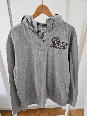Grå hoodie från Massif camp  - Snygg grå hoodie från Massif camp med tryck och broderi på bröstet. Hoodien har knappar vid halsen, huva och långa ärmar. Tillverkad i mjukt bomullsmaterial som känns skönt mot huden. Perfekt för en avslappnad och sportig stil.