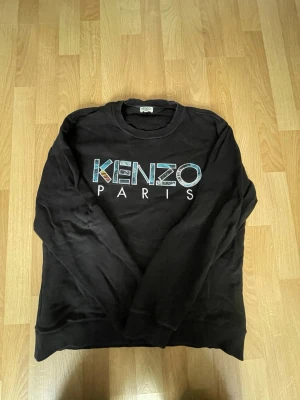 Svart sweatshirt från Kenzo Paris - Cool svart sweatshirt från Kenzo Paris med stort färgglatt logotryck på bröstet. Klassisk rund halsringning och långa ärmar. Tillverkad i mjukt material som känns skönt mot huden. Perfekt för en avslappnad och trendig look.