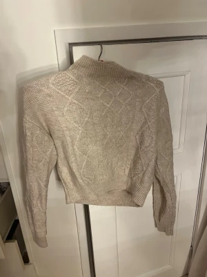 Beige stickad tröja med mönster - Säljer en beige stickad tröja med höghals och snyggt kabelstickat mönster på både ärmar och rygg. Tröjan har en croppad passform och ribbade muddar vid hals, ärmslut och nederkant. Perfekt för dig som gillar en stilren och mysig look. 