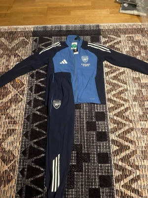 Arsenal träningsdress från Adidas - Snygg Arsenal träningsdress från Adidas i blått och marinblått. Jackan har dragkedja, vita Adidas-ränder på axlarna och Arsenal-logga på bröstet. Byxorna har smal passform, vita ränder och Arsenal-märke på låret. Perfekt för fotbollsträning eller chill.