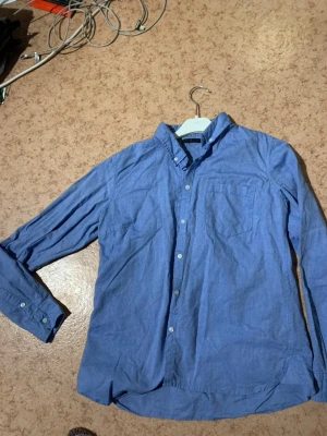 Blå bomullsskjorta med knappar - Klassisk blå skjorta i bomull med lång ärm och knäppning framtill. Skjortan har en bröstficka och button down-krage för en clean look. Perfekt att styla lager på lager eller bära som den är.