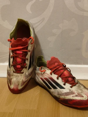 Adidas vita och röda fotbollsskor - Säljer ett par Adidas fotbollsskor med vit bas, röda detaljer och svarta ränder på sidorna. Skorna har snörning med röda skosnören och en låg profil. Ovandelen är tillverkad i syntetmaterial och designen är sportig och modern, perfekt för snabba spelare på planen.