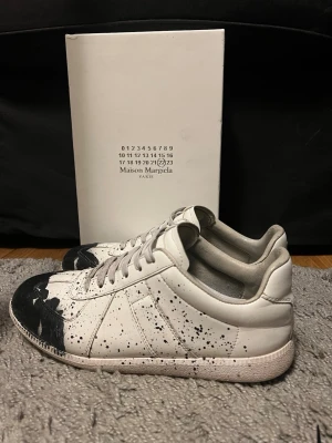 Maison Margiela vita sneakers med svart färgstänk - Maison Margiela sneakers i vitt läder med svart färgstänk och målad tå. Klassisk låg modell med vita snören och gummisula. Unik design med konstnärlig touch, perfekt för dig som gillar statement-plagg. Skick 8,5/10