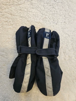 Mörkblå tumvantar Polarn O. Pyret 1till2 år - Mörkblå tumvantar från Polarn O. Pyret med reflexrand framtill och justerbart band runt handleden. Vantarna har greppvänlig yta i handflatan och är perfekta för kalla dagar.