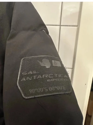 Svart dunjacka Sail Racing - Svart dunjacka från Sail Racing med broderad patch på ärmen och logga på bröstet. Jackan har huva, dragkedja framtill och två bröstfickor med dragkedja. Perfekt för kalla vinterdagar och inspirerad av Antarktis-expedition.