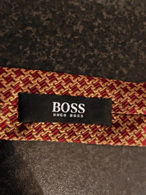 Mönstrad slips från Hugo Boss - Säljer en snygg slips från Hugo Boss med ett tätt mönster i vinrött och guld. Slipsen har en klassisk form och känns riktigt lyxig tack vare sitt material och design. Perfekt för dig som vill sticka ut med stilrena detaljer.