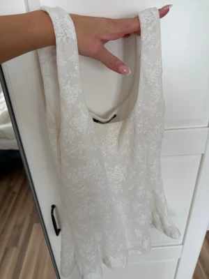 Vit spetslinne med blommönster - Snyggt vitt linne med breda axelband och blommigt spetsmönster. Linnet har en lätt utsvängd passform och är perfekt för varma dagar. Materialet känns lätt och luftigt, och spetsen ger en romantisk vibe.