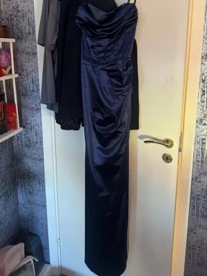 Mörkblå lång satinklänning - Säljer en elegant mörkblå långklänning i glansigt satintyg. Klänningen har tunna axelband, draperad byst och snörning framtill för en snygg passform. Perfekt för dig som vill ha en stilren och trendig look. Lik den som finns på Bubbelroom 