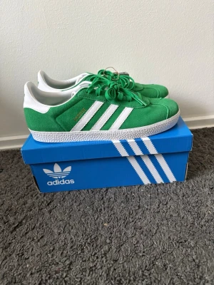 Adidas Gazelle gröna sneakers - Gröna Adidas Gazelle sneakers i mocka med klassiska vita ränder på sidorna och vit hälkappa. Skorna har platt sula och snörning framtill. Guldtext med modellnamn på sidan och ikonisk retrostil. Perfekta för dig som gillar streetwear och sportig look.