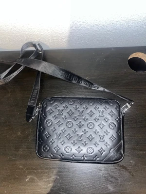 Lv väska - Snygg svart axelväska från Louis Vuitton i präglat läder med klassiskt monogrammönster. Väskan har justerbar axelrem med Louis Vuitton-tryck och silverfärgade detaljer. Perfekt för dig som vill ha en stilren och lyxig accessoar. Kom med bud!
