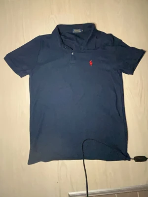 Marinblå pikétröja från Polo Ralph Lauren - Klassisk marinblå pikétröja från Polo Ralph Lauren med korta ärmar och krage. Tröjan har den ikoniska röda logotypen broderad på bröstet och knäppning vid halsen. Perfekt till jeans eller shorts för en clean och stilren look.Står storlek M men skulle säga det är en S ungefär. Hör av dig om minsta fundering!