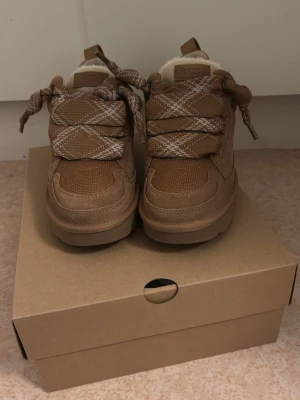 Bruna UGG sneakers med ullfoder - Snygga bruna sneakers från UGG med beige och vita mönstrade skosnören. Ovandelen är i mocka och mesh, och insidan har mjukt ullfoder. Skorna har en chunky sula och låg profil, perfekt för dig som gillar streetwear och vill ha något extra bekvämt.
