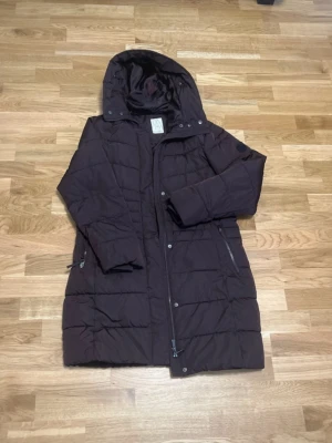 Mörkbrun dunjacka från Etirel - Snygg mörkbrun dunjacka från Etirel med huva och dragkedja framtill. Jackan har två sidofickor med dragkedja och quiltad design som ger en modern look. Perfekt för kalla vinterdagar och riktigt skön att bära.