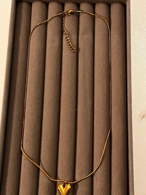 Guldigt halsband med hjärthänge - Snyggt halsband i guldfärgad metall med ett blankt hjärthänge. Kedjan är tunn och elegant med justerbar längd och karbinlås. Hjärtat har en modern, lite droppformad design som verkligen sticker ut.
