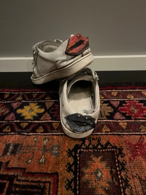 Vita sneakers med läppdetalj bak - Unika vita sneakers i skinn med snörning och coola läppar i blått och rött bak på hälen. Skorna har platt sula och rund tå, och detaljer i metall på läpparna. Perfekt för dig som vill sticka ut med din stil.