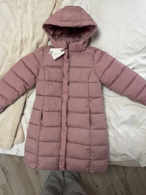 Ljusrosa lång dunjacka med huva - Säljer en ljusrosa lång barn dunjacka från 157 med huva och quiltad design. Jackan har dragkedja och tryckknappar framtill samt två sidofickor. Perfekt för kalla vinterdagar och riktigt skön att bära. Materialet är mjukt och värmande.