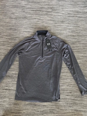 Grå långärmad träningströja UA - Snygg grå långärmad träningströja från Under Armour med halv dragkedja framtill och hög krage. Tillverkad i ett mjukt och stretchigt funktionsmaterial som andas. Perfekt för löpning eller gym. UA-logga på bröstet och modern passform.