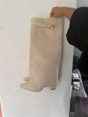 Beige mockastövlar  - Snygga beige stövlar i mocka med spetsig tå och bred klack. Dekorativt spänne med gulddetalj upptill. Stövlarna har en stilren och lyxig look som passar perfekt till höstens och vinterns outfits. Helt nya och oanvända! Storlek 39 men passar även på mig som har 38! 