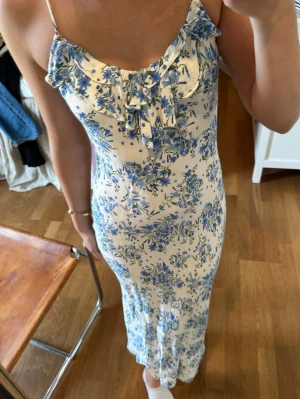 Blommig långklänning med volang - Långklänning med smala axelband i ljus bottenfärg och blått blommönster. Klänningen har volangdetaljer vid urringningen och en feminin, figurnära passform. Perfekt för sommarens alla tillfällen.