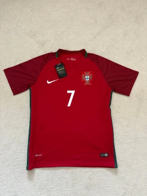 Portugal  - Säljer en röd Portugal fotbollströja från Nike med nummer 7 och Ronaldo på ryggen. Tröjan har korta ärmar, gröna detaljer längs sidorna och är tillverkad i Dri-FIT material som andas. Perfekt för dig som älskar fotboll och vill stötta Portugal. Helt ny och oanvänd