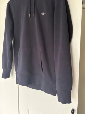 Mörkblå hoodie från GANT - Snygg mörkblå hoodie från GANT med liten broderad logga på bröstet. Klassisk känguruficka framtill, ribbade muddar och dragsko i huvan. Tillverkad i mjuk bomullsblandning för skön känsla. Perfekt för en avslappnad och stilren look.