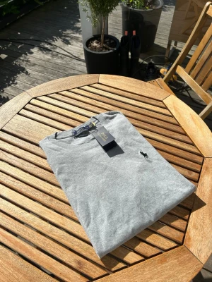 Grå t-shirt från Polo Ralph Lauren - Snygg grå t-shirt från Polo Ralph Lauren med klassisk svart logga broderad på bröstet. T-shirten har rund hals och är tillverkad i mjuk bomull för en skön känsla. Perfekt basplagg för alla tillfällen.