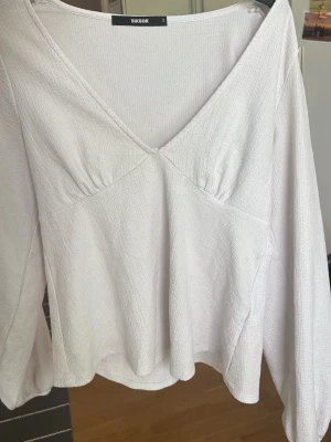 Vit v-ringad blus från Bik Bok - Säljer en vit topp från Bik Bok med v-ringning och långa ärmar. Toppen har en lätt struktur i tyget och en avslappnad passform. Perfekt att styla med jeans eller kjol för en enkel och fräsch look.