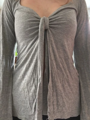 Designbysi maddytop - Trendig grå långärmad topp med knyt framtill och lätt v-ringad hals. Toppen har en croppad känsla och är tillverkad i mjukt bomullsmaterial. Perfekt för dig som gillar en avslappnad men ändå snygg stil.