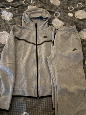 Grått Nike loungewear set M - Snyggt grått loungewear set från Nike med zip-hoodie och matchande byxor. Setet har svarta detaljer och Nike-logga på både tröja och byxa. Tillverkat i mjukt material, perfekt för chill dagar hemma eller när du vill vara bekväm.