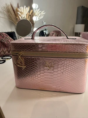 Rosa sminkväska i ormmönstrat material - Säljer en glansig rosa sminkväska från Victoria secret med ormmönstrad yta. Väskan har ett praktiskt handtag, guldfärgad dragkedja och hjärtformad detalj. Perfekt för att organisera smink eller accessoarer.