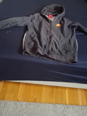 Svart Nike Air hoodie med dragkedja - Svart hoodie från Nike Air med dragkedja framtill och huva. Mjuk fleece på insidan för extra komfort. Nike Air-logga i orange på bröstet och två fickor på sidorna. Perfekt för chill dagar eller när du vill ha en sportig look.