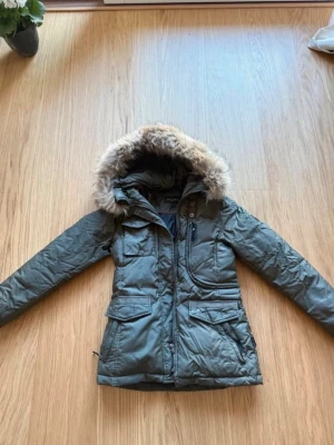 Mörkgrön parkas med fuskpäls från Hollies  - Snygg mörkgrön parkas från Hollies med stor huva och fuskpäls. Jackan har flera praktiska fickor, dragkedja och knappar framtill samt en patch på ärmen. Perfekt för kalla dagar och ger en cool streetwear-look.