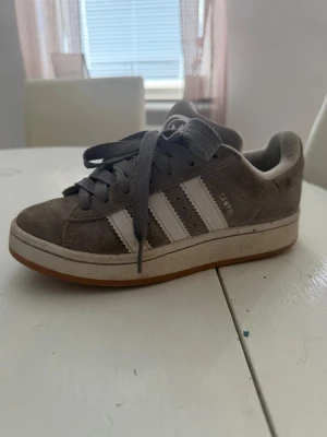 Adidas Campus grå sneakers - Gråa Adidas Campus sneakers i mocka med klassiska vita ränder på sidorna och gummisula. Skon har låga skaft, gråa skosnören och Adidas logga på plösen. Perfekt för en avslappnad streetwear-look. Rengörs sjävklart innan försäljningen 