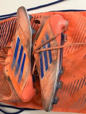 Adidas f50 - Säljer ett par snygga orangea Adidas f50 elite fotbollsskor med blåa detaljer och klassiska tre ränder på sidan. Skorna har snörning. Ovandelen är i syntetmaterial och designen är riktigt sportig och modern.