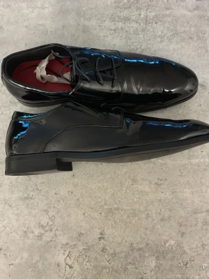 Svarta lackskor med snörning - Snygga svarta lackskor med klassisk snörning och låg klack. Skorna har en glansig finish och röd insida, perfekt för dig som vill sticka ut med en stilren look. Passar till kostym eller när du vill vara extra uppklädd. Box ingår och dem är köpte i Butiken VOLT. Ny pris är 1800.
