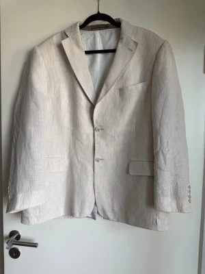 Beige randig kavaj i linneblandning - Snygg beige kavaj med tunna vita ränder och klassisk passform. Kavajen har två knappar framtill, fyra dekorativa knappar vid ärmslut och två fickor med lock. Tillverkad i en luftig linneblandning som passar perfekt till sommaren.