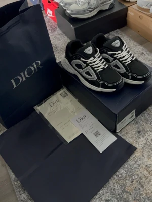 Dior b30 svart/vit - Säljer ett par svarta Dior B30 sneakers med vita och grå detaljer. Skorna har chunky sula, vita skosnören och en modern, sportig design. Tillverkade i en mix av mesh och läder för både stil och komfort. Perfekta för dig som vill sticka ut med exklusiva sneakers.