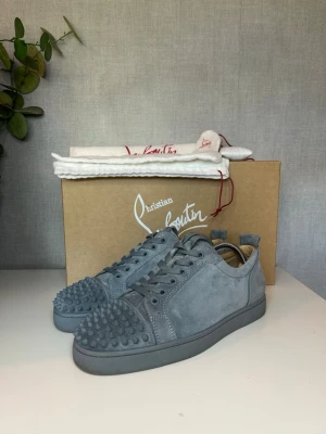Gråa Christian Louboutin sneakers - Snygga gråa sneakers från Christian Louboutin i mocka med ikoniska nitar på tån. Skorna har en låg silhuett, matchande grå snörning och en klassisk gummisula. Perfekta för dig som vill sticka ut med exklusiv design och edgy detaljer. 40 1/2 passar 41 1/2 