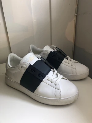 Valentino vita sneakers med svart band - Är lite mer mörkblåa i verkligheten!Snygga vita sneakers från Valentino med ett brett svart band över vristen och vita skosnören. Skorna har en stilren design med guldfärgad logga på plösen och är tillverkade i slätt läder. Perfekta för dig som gillar exklusiva och trendiga detaljer.