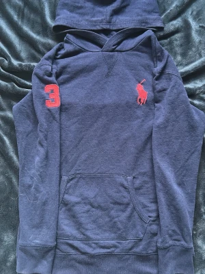Marinblå hoodie från Polo Ralph Lauren - Snygg marinblå hoodie från Polo Ralph Lauren med röd logga på bröstet och en röd siffra 3 på ärmen. Klassisk känguruficka framtill och huva. Tillverkad i mjuk bomull för skön känsla och avslappnad stil.